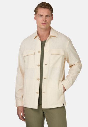 Giacca beige chiaro realizzata in tessuto, con chiusura a bottoni, due tasche sul petto e colletto. Indossata sopra una camicia verde.