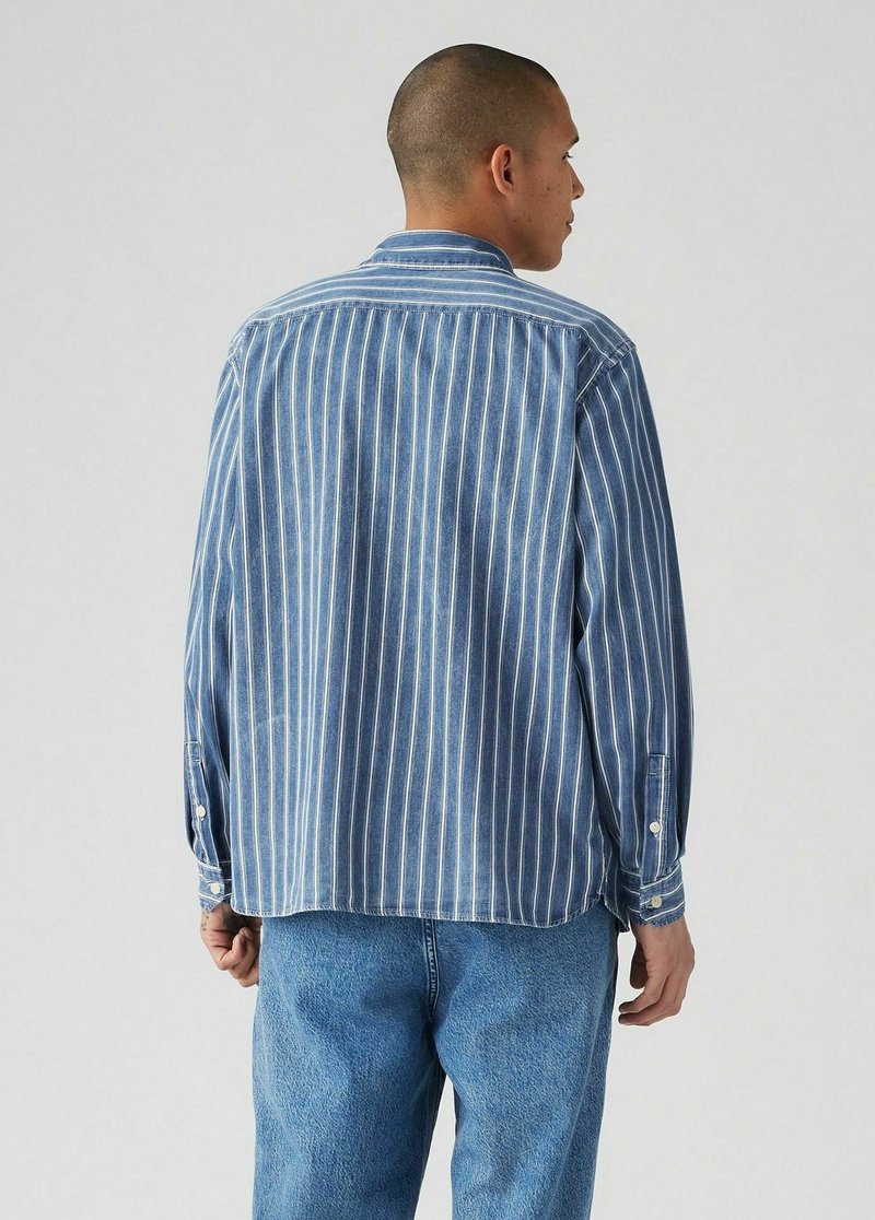 Chemise en denim à rayures bleues avec des manches longues, rayures verticales bleu clair et blanches, poignets à boutons et col classique, associée à un jean bleu clair.