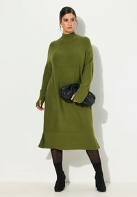 Robe en tricot vert avec un col haut, des poignets côtelés et un ourlet. Associée à un sac noir et des bottines noires. Texture lisse, coupe décontractée.