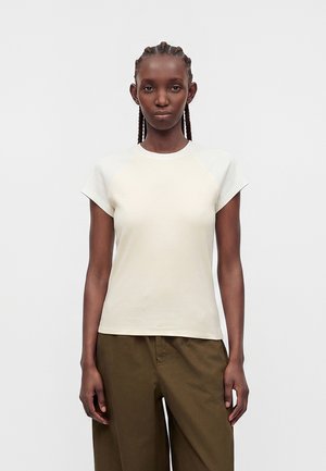 Jeune personne aux cheveux tressés portant un t-shirt blanc uni à manches courtes et un pantalon ample vert olive foncé, debout devant un fond blanc.