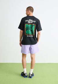 Sort grafisk t-shirt med et grønt naturdesign og teksten "Big Bend, TEXAS" på ryggen, parret med lyse lilla shorts og sneakers.