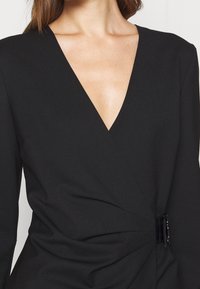 Schwarzes Wickelkleid mit tiefem V-Ausschnitt, strukturiertem Stoff und seitlichem Raffungselement, das mit einem glänzenden schwarzen Akzent gesichert ist.