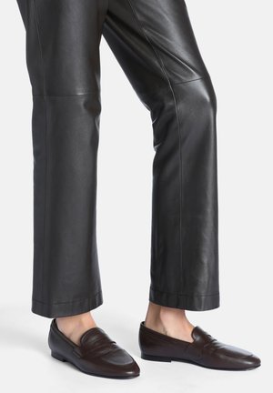 Personne portant un pantalon en cuir noir et des mocassins en cuir marron foncé sans lacets avec des petits talons sur fond blanc.