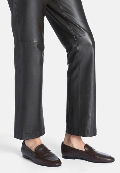 Personne portant un pantalon en cuir noir et des mocassins en cuir marron foncé sans lacets avec des petits talons sur fond blanc.