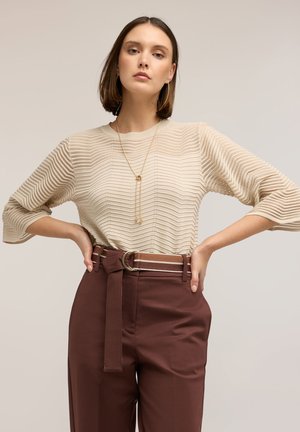 Donna con maglione beige dalla texture lavorata, pantaloni marroni a vita alta con cintura abbinata e lunga collana d'oro, in piedi con le mani sui fianchi.