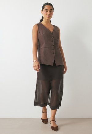 Gilet sans manches marron avec fermeture à boutons, assorti à une jupe noire transparente. Texture suédée, escarpins à bout pointu avec brides de cheville.