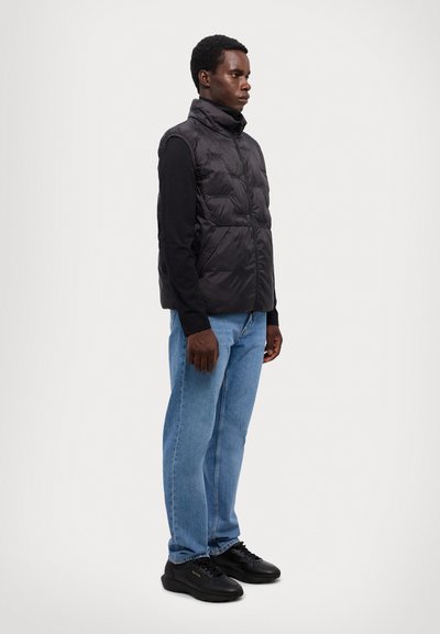 Sort polstret vest med høj krave over en langærmet sort skjorte, parret med lyseblå jeans og sorte sneakers. Glat tekstur, lynlås lukning.
