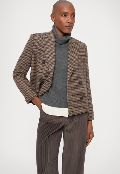 Brązowa marynarka w kratkę houndstooth z podwójnym zapięciem i czarnymi guzikami, szary golf, beżowa koszula jako warstwa, oraz teksturowane brązowe spodnie z kordurowej tkaniny.