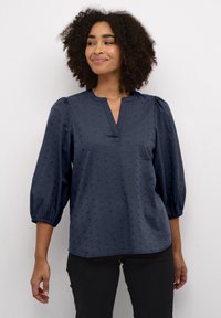 Blouse bleu marine avec un décolleté en V, des manches bouffantes trois quarts et un motif en polka dots texturé. Le matériau semble léger et doux.