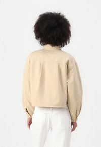 Jaqueta cropped bege com um tecido texturizado, gola e punhos elásticos, acompanhada de jeans brancos, apresentando um corte relaxado.