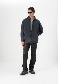 Jaqueta de denim preta com fecho na frente, apresenta uma gola alta e bolsos laterais. Combinada com calças de imitação de pele pretas e ténis elegantes.
