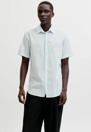 Homme portant une chemise bleu clair à manches courtes avec un petit motif à pois et un pantalon noir, debout devant un fond uni.