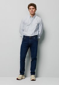 Lichtblauw en wit gestreept overhemde, donkere denim jeans en beige sneakers. Heeft een klein logo op de borst van het shirt.