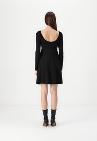 Vestido preto de manga longa com corpete justo e saia rodada, apresentando uma profunda abertura nas costas e uma textura suave e macia. Usado com botas pretas até os tornozelos.