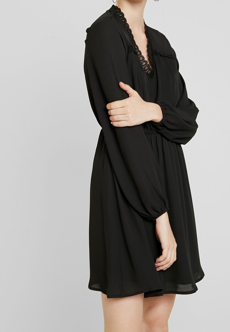 Svart långärmad klänning med djup V-ringning, featuring spetsdetaljer, elastisk midja och en flödande kjol som når ovanför knäna.