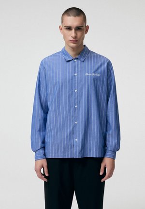 Młody mężczyzna w niebieskiej, długorękawowej koszuli zapinanej na guziki z białymi pionowymi paskami i ciemnych spodniach, stojący na tle jasnego, jednolitego tła.