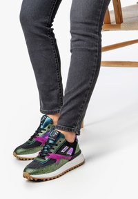 Henkilö, jolla on päällään mustat farkut ja värikkäät sneakersit, joissa on vihreitä, purppuran värisiä ja mustia kuvioita, istuu puupenkissä, jossa on punottu istuin.