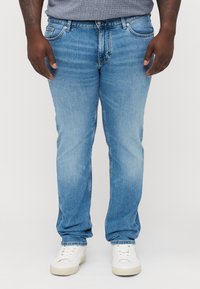 Mężczyzna ubrany w jasnoniebieskie dżinsy, szarą koszulę, białe trampki, stoi z rękami swobodnie opuszczonymi wzdłuż ciała na jednolitym tle.