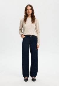 Gaisīgi bēšs, garām piedurknēm adīts džemperis ar mīkstu tekstūru, kopā ar tumši zilām, plašām džinsu biksēm, kurām ir klasisks džinsa dizains un jostas cilpas.