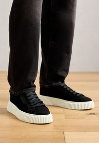 Person wearing black suede sneakers with thick white ribbed soles and black laces, standing on light wood floor.