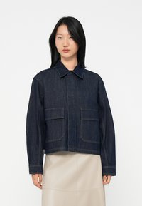 Kvinne med rett svart hår iført en mørkeblå denimjakke med store frontlommer og en beige skjørt, som står mot en hvit bakgrunn.