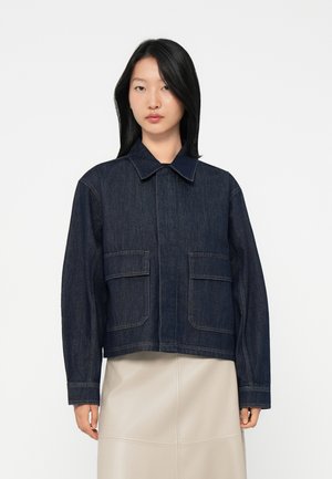 Kvinne med rett svart hår iført en mørkeblå denimjakke med store frontlommer og en beige skjørt, som står mot en hvit bakgrunn.