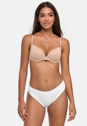 Woman with long dark hair wearing a nude bra and white underwear, standing against a plain light background.