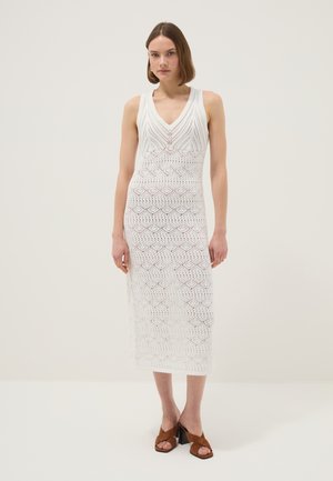Frau trägt ein ärmelloses weißes Häkel-Midikleid mit V-Ausschnitt und braunen Sandalen mit Absatz, steht vor einem schlichten hellen Hintergrund.