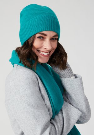 Gorro de punto en teal y bufanda a juego, hechos de un tejido suave. La bufanda cae sobre un abrigo gris claro. Se puede ver la textura a rayas en el gorro.