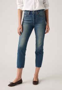 Raka blå denimjeans med hög midja, ljus blekning och fransade fållar. Matchas med bruna platta skor och en vit skjorta med knappar.
