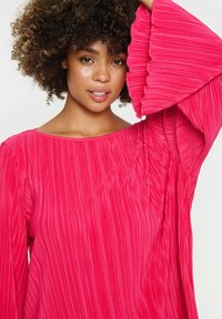 Zračna roza bluzica s okroglim izrezom in dolgimi, volančastimi rokavi; teksturirana tkanina prikazuje navpične naborke.