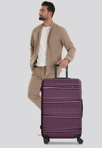 Hombre con un traje color beige de pie, con una mano en el bolsillo y la otra sosteniendo el asa de una gran maleta rígida morada con ruedas.