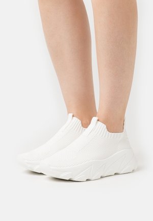 Ben iført hvide slip-on strikkede sneakers med tykke såler, stående på en ensfarvet hvid baggrund.