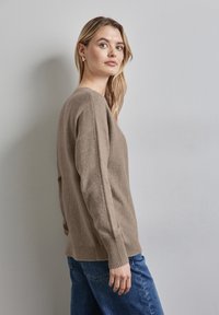 Hellbrauner, gestrickter Pullover mit rundem Ausschnitt und gerippten Bündchen, kombiniert mit blauen Jeans. Der Stoff wirkt weich und strukturiert.