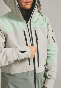 Lightweight, color-blocked jacket with mint green and gray panels, featuring a hood, black zippers, and adjustable cuffs. Smooth texture.