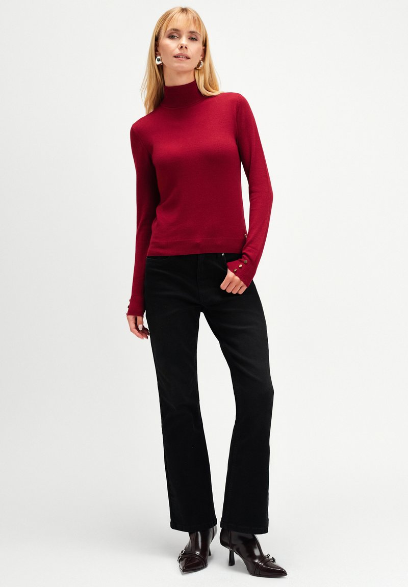 Kobieta w dopasowanym czerwonym swetrze z golfem i detalami w postaci guzików, czarnych prostych spodniach oraz ciemnych brązowych botkach na obcasie.