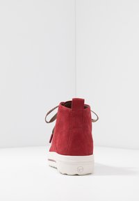 Czerwona zamszowa botka za kostkę z zaokrąglonym noskiem, brązowymi sznurowadłami i białą gumową podeszwą. Posiada pętelkę z tyłu oraz ozdobne przeszycia.