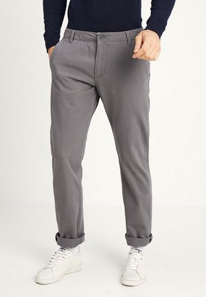 Mann trägt graue hochgekrempelte Hose, weiße Sneakers und ein schwarzes Langarmshirt, steht mit einer Hand in der Tasche.