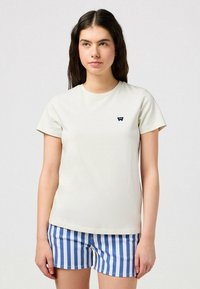 Camiseta de algodón color crema claro con mangas cortas, cuello redondo y un pequeño logotipo azul oscuro en el pecho. Combinada con pantalones cortos a rayas en azul y blanco.