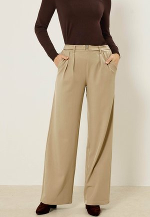 Kvinde iført beige vidde bukser med plisseringer og sidelommer, kombineret med en brun langærmet top og mørkebrune sko.