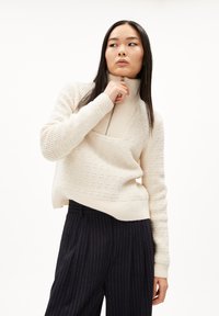 Maglione a collo alto testurizzato color crema con zip a metà e polsini a coste, abbinato a pantaloni navy a righe.