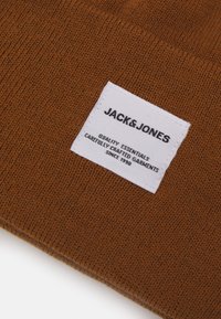 Gorro de punto marrón con una etiqueta blanca. La etiqueta presenta "JACK & JONES" en texto negro y texto adicional en blanco sobre la calidad.
