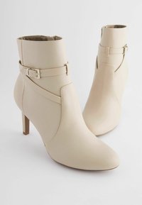 Stivali alla caviglia beige realizzati in pelle liscia con punta affusolata, tacco alto e sottile, e cinturino decorativo intorno alla caviglia.