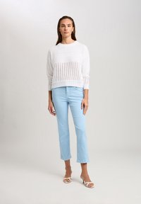 Weißer gestrickter Pullover mit strukturiertem Muster, kombiniert mit hellblauen, figurbetonten Hosen. Das Model steht in weißen Riemchensandalen.