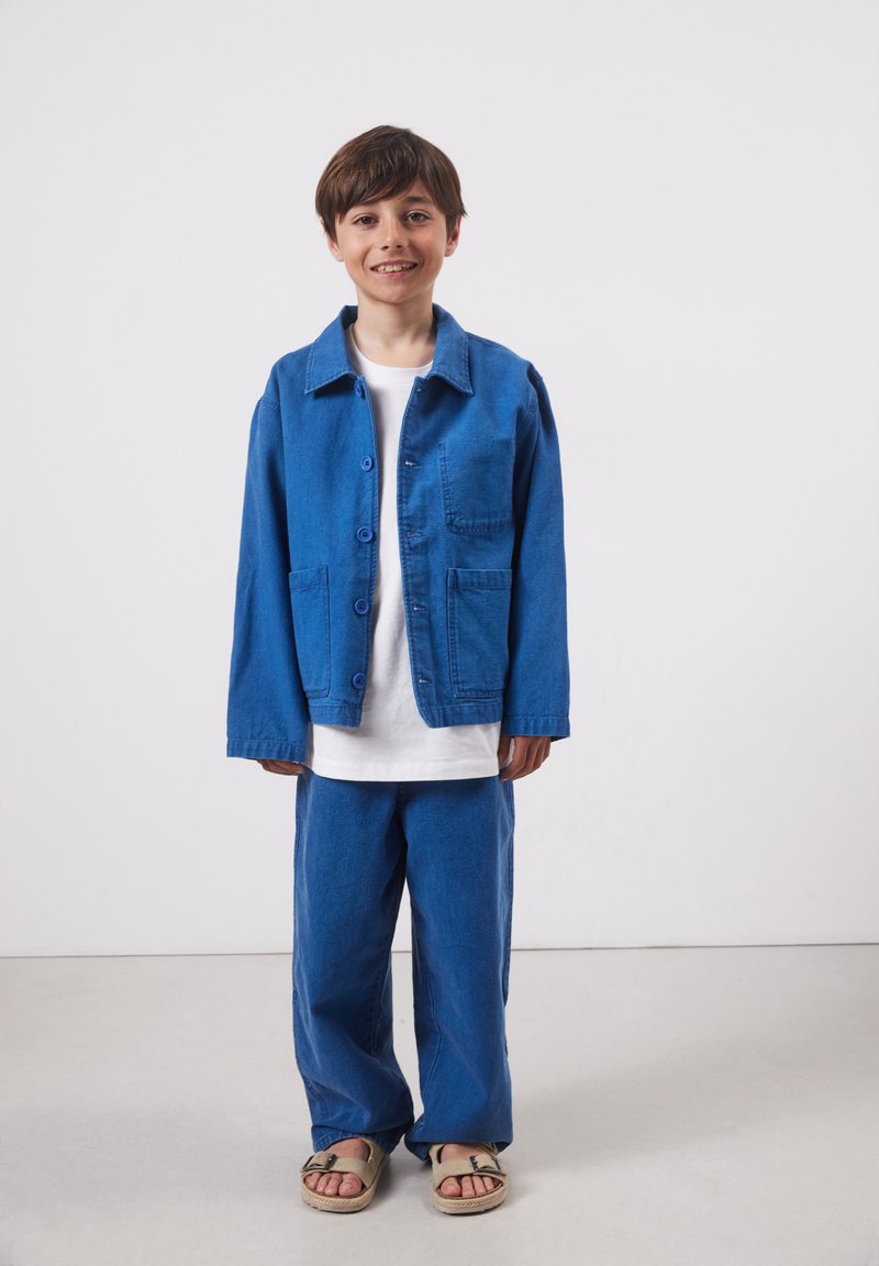 Garçon debout, souriant, portant une veste boutonnée bleue, un pantalon assorti, un t-shirt blanc et des sandales beiges sur un fond clair.