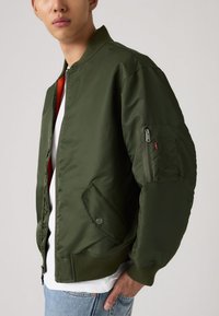 Mann iført olivengrønn bomberjakke med glidelåslomme på ermet, fremre klafflommer, hvit skjorte og lyse blå jeans, med hånden i lomma.