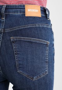 Jeans de mezclilla azul oscuro con un parche de cuero etiquetado como "WEEKDAY" en la cinturilla y dos bolsillos traseros con costuras contrastantes.