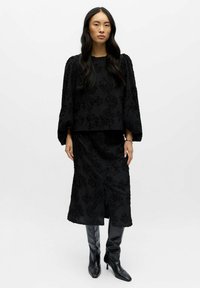 Moteris su ilgais juodais plaukais, dėvinti juodą tekstūruotą gėlių midi sijoną ir atitinkamą ilgomis rankovėmis bluzoną, su juodais iki kelių batais.