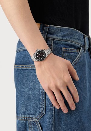 Montre en acier inoxydable argenté avec un cadran noir, des index horaires blancs et des accents rouges. Portée au poignet, associée à un jean en denim bleu.
