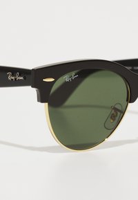 Ochelari de soare negri Ray-Ban cu lentile verzi, având o ramă din metal cu tonuri de aur, design pătrat îndrăzneț și logo-ul mărcii pe rame.